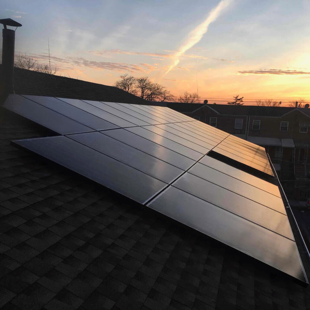 A rooftop solar panel installation at sunset, reflecting the skys orange and pink hues. Scattered clouds dot the horizon, with silhouettes of buildings and trees in the background—an ideal scene for contemplating battery backup installation.