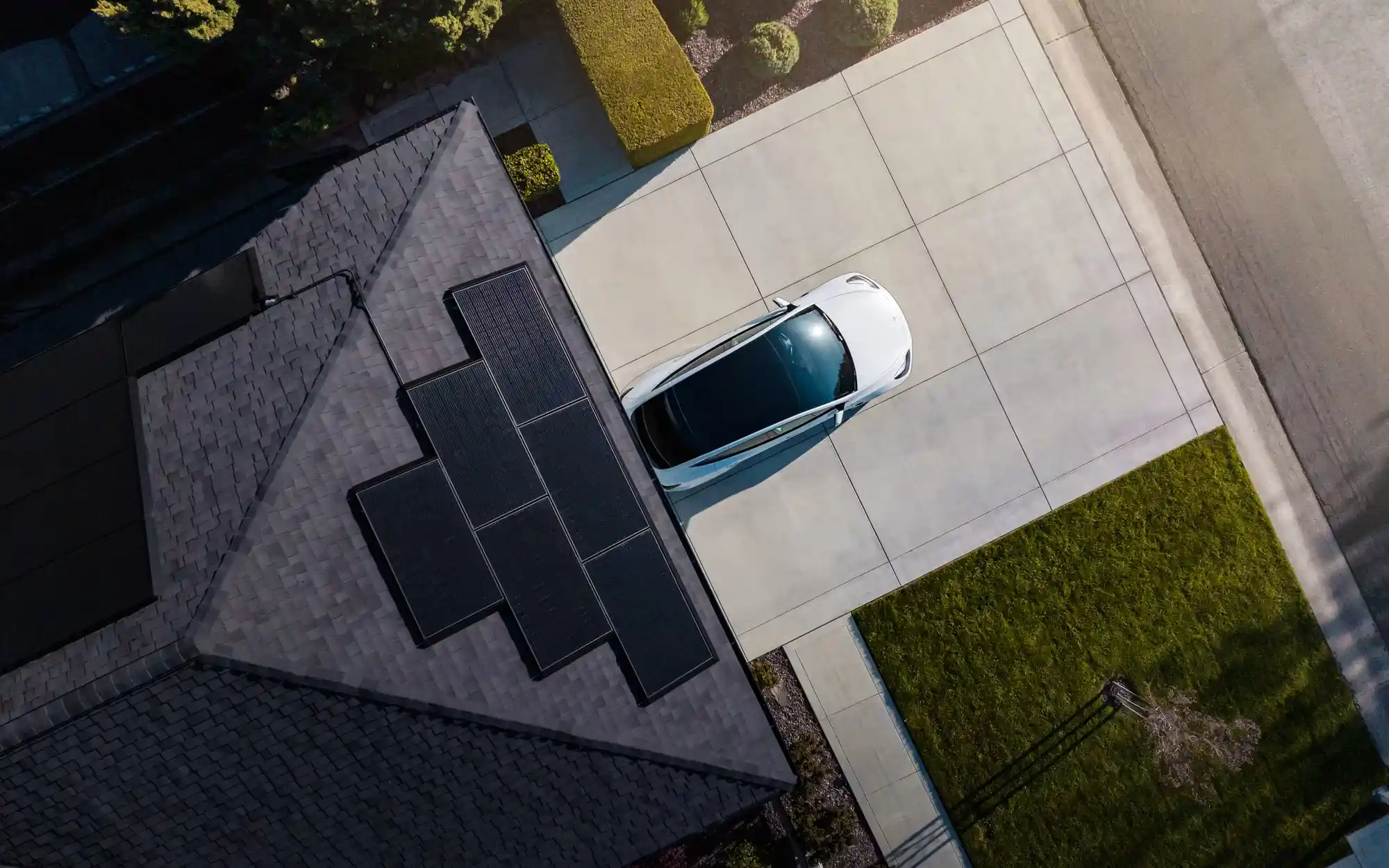 Aerial view of a house boasting solar panel installation on the sunlit roof. A white car rests in the driveway, surrounded by a lush green lawn and pavement. The scene is vividly illuminated by sunlight.