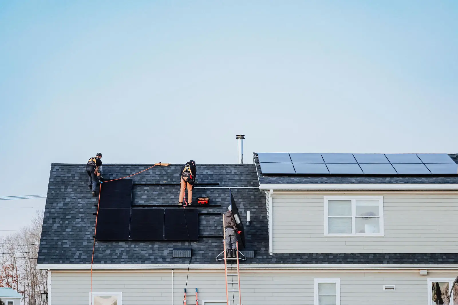 Three workers install black solar panels on a light gray house, while smart electrical panels manage energy from other solar panels already in place on an adjacent roof section.