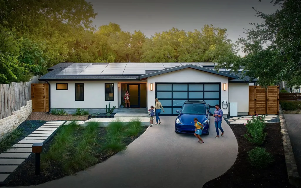 Situated in Massachusetts, this modern home boasts solar panels on the roof and is nestled among lush trees. A family of four enjoys the outdoors as their blue electric car sits in the driveway. Children play while adults watch, with the homes exterior showcasing a blend of white and dark panels.