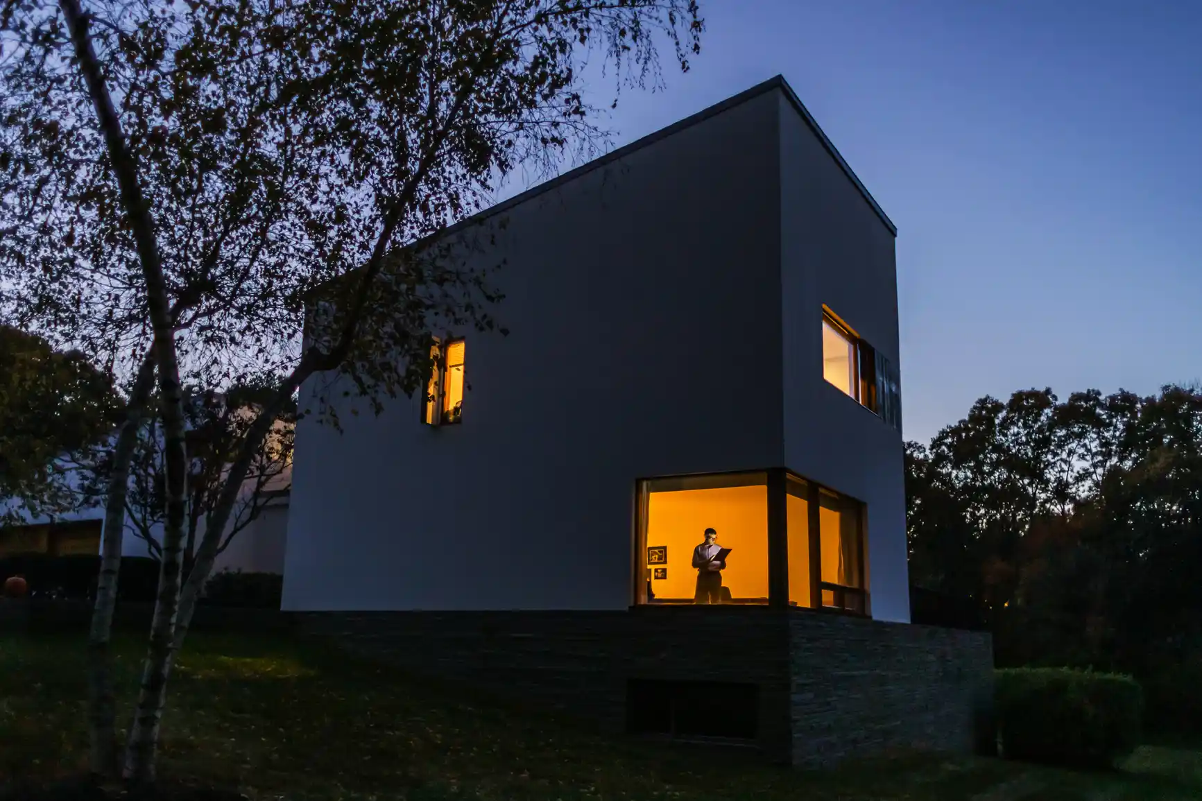 A modern, white house with large windows is illuminated from within at dusk, thanks to solar power. A person is visible inside on the ground floor, casting a warm glow.